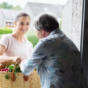 istockphoto-944961100-612×612-1 Young woman delivers groceries, mainly food such as vegetables, bread and mild in a shopping bag, to a senior woman. Senior woman standing at open door of her house, happy to receive the support of community outreach